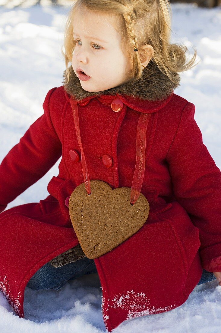 Small girl with gingerbread heart sitting in snow