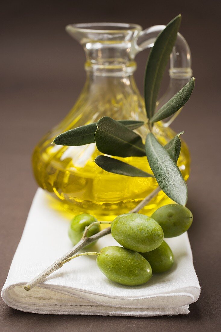 Grüne Oliven am Zweig, dahinter Olivenöl in Karaffe