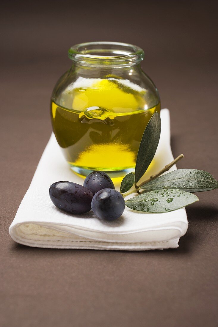 Schwarze Oliven am Zweig, dahinter Olivenöl im Glas