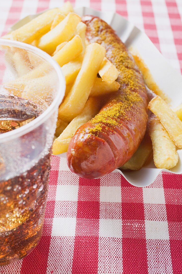 Currywurst mit Pommes frites auf Pappteller, Cola im Becher