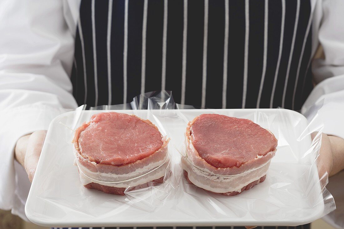 Person holding two bacon-wrapped beef medallions on tray