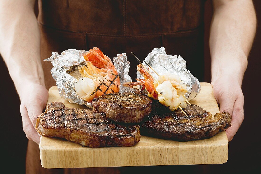 Man holding Surf & Turf (steaks & prawns) on chopping board