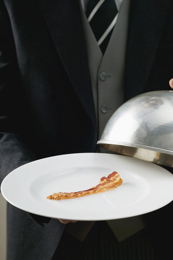Butler serving rasher of fried bacon on plate with dome cover