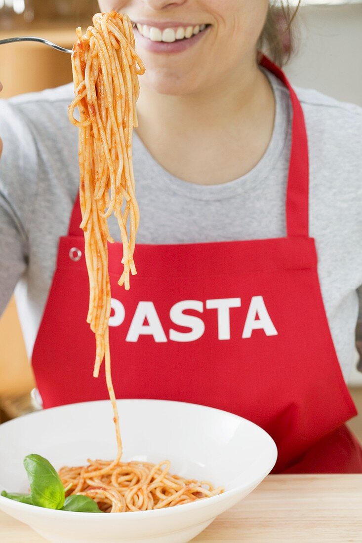 Young woman in apron eating spaghetti with tomato sauce