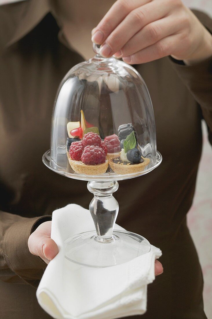 Woman holding assorted berry tarts under glass serving dome