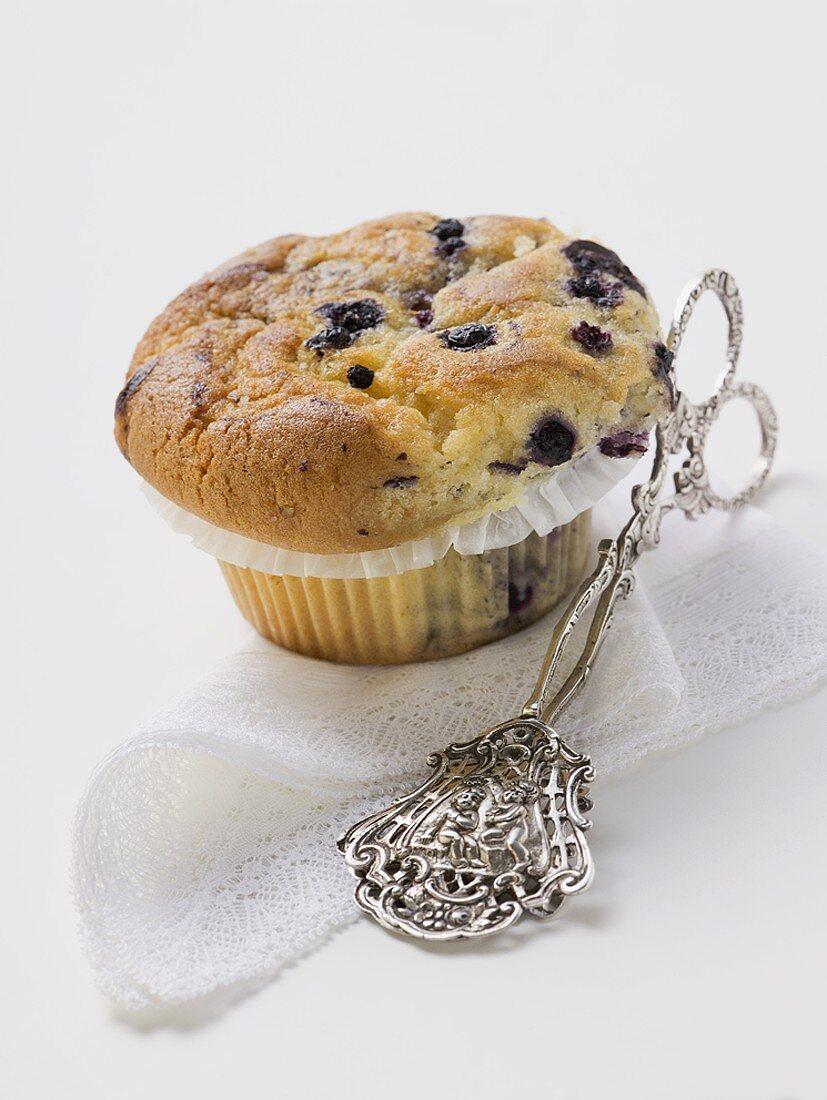 Blueberry muffin in paper case, cake tongs beside it