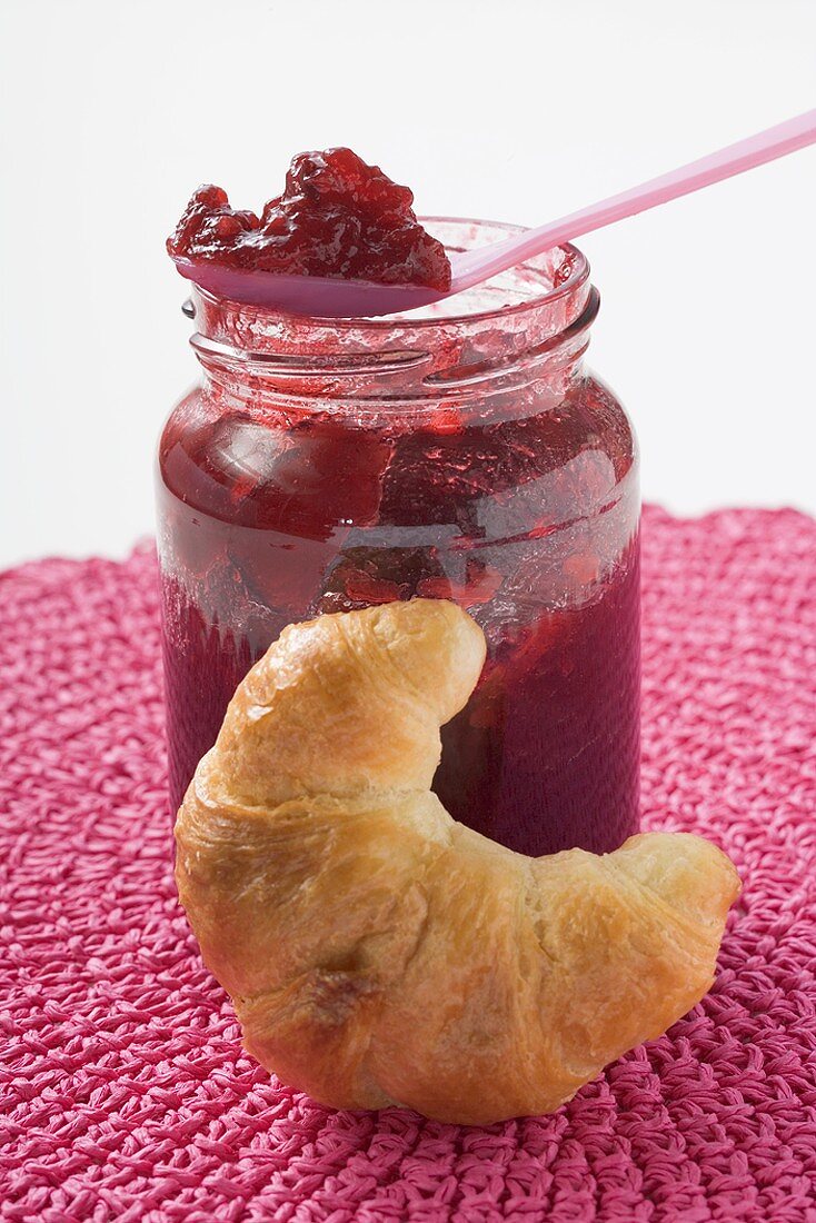 Jar of raspberry jam with spoon, croissant in foreground