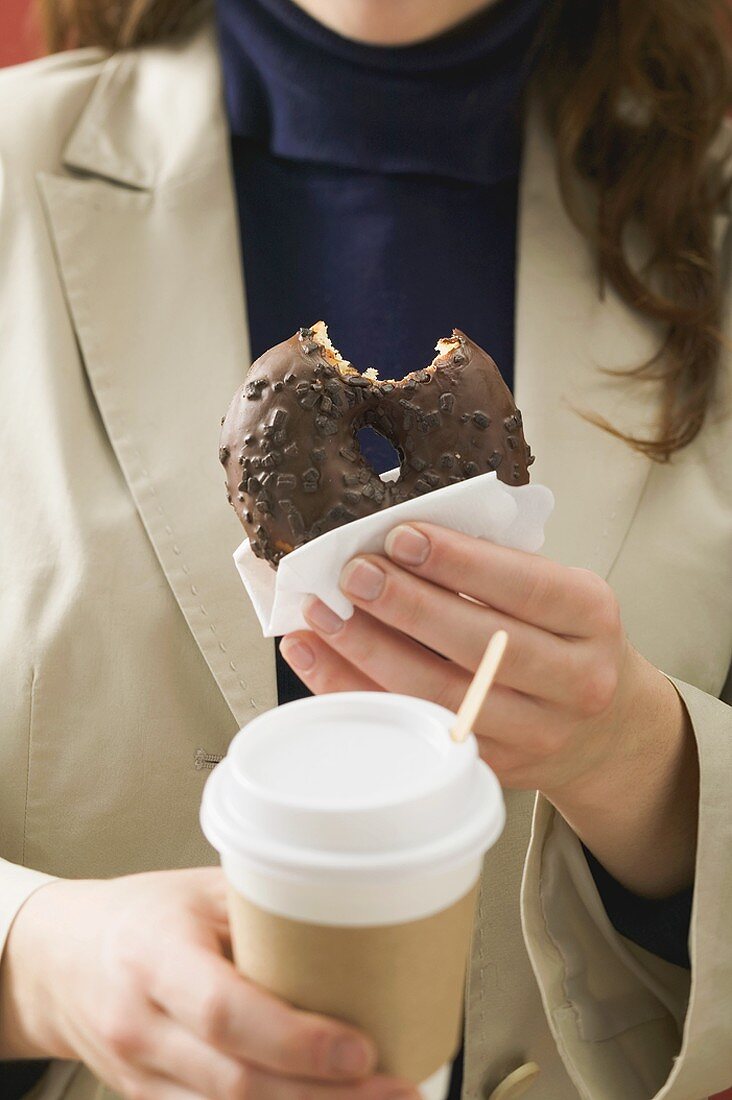 Woman holding chocolate doughnut with bite taken & cup of coffee