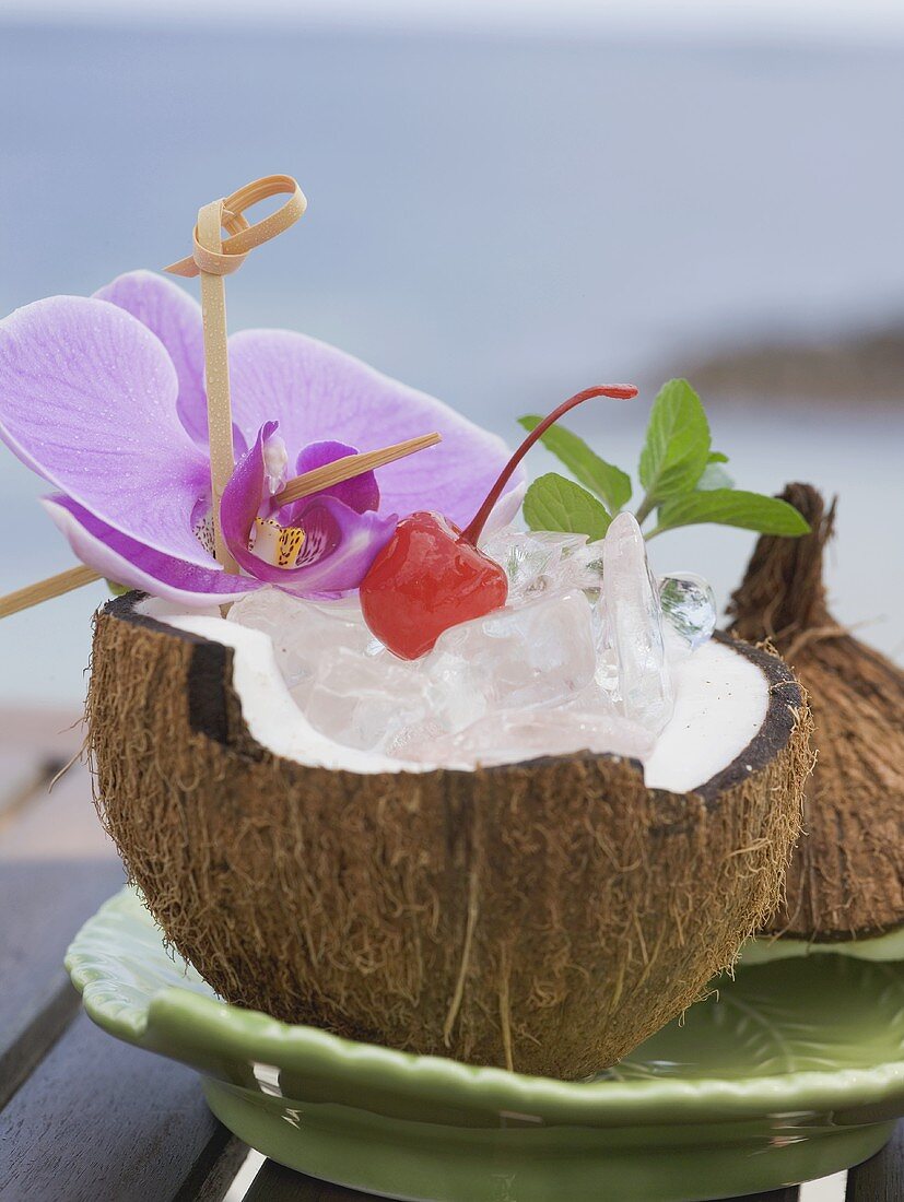Kokosnusshälfte mit Eiswürfeln, Cocktailkirsche und Orchidee