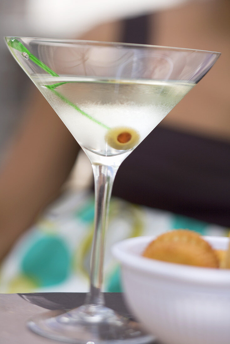 Martini with green olive, crackers, woman in background