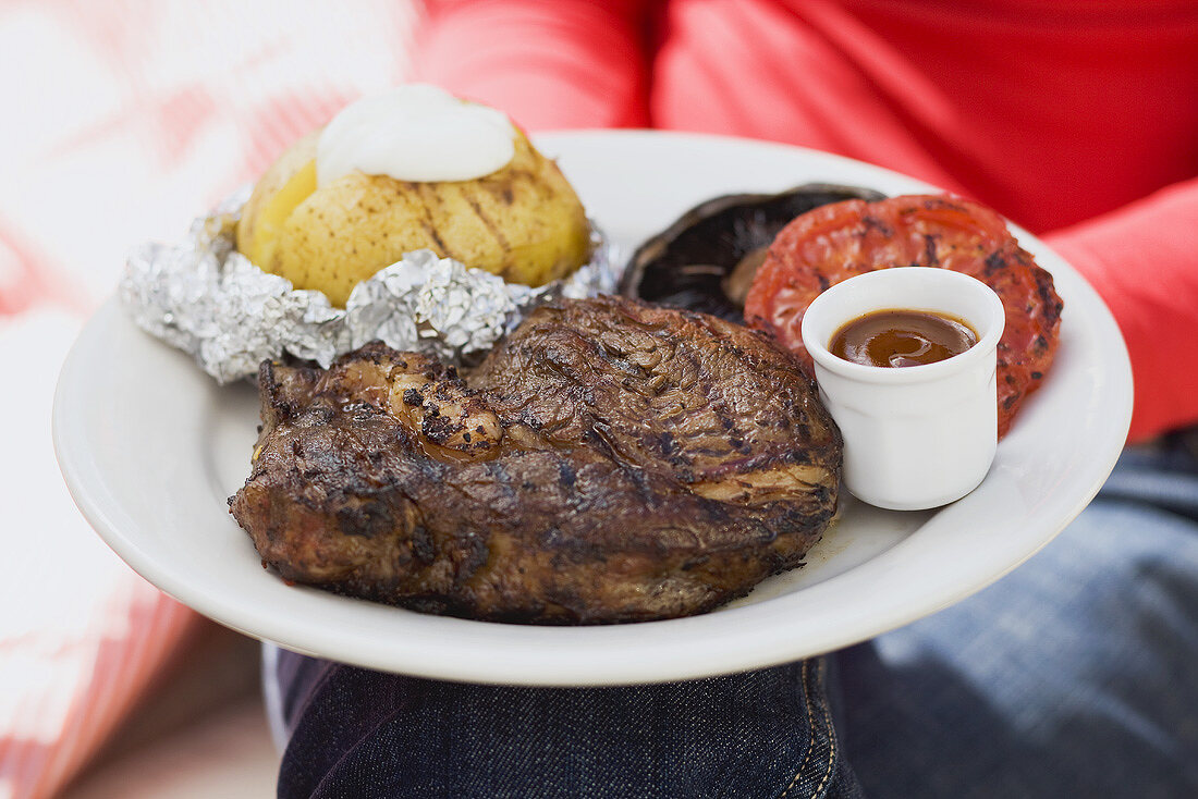 Frau hält Teller mit Steak, Folienkartoffel, Gemüse und Sauce