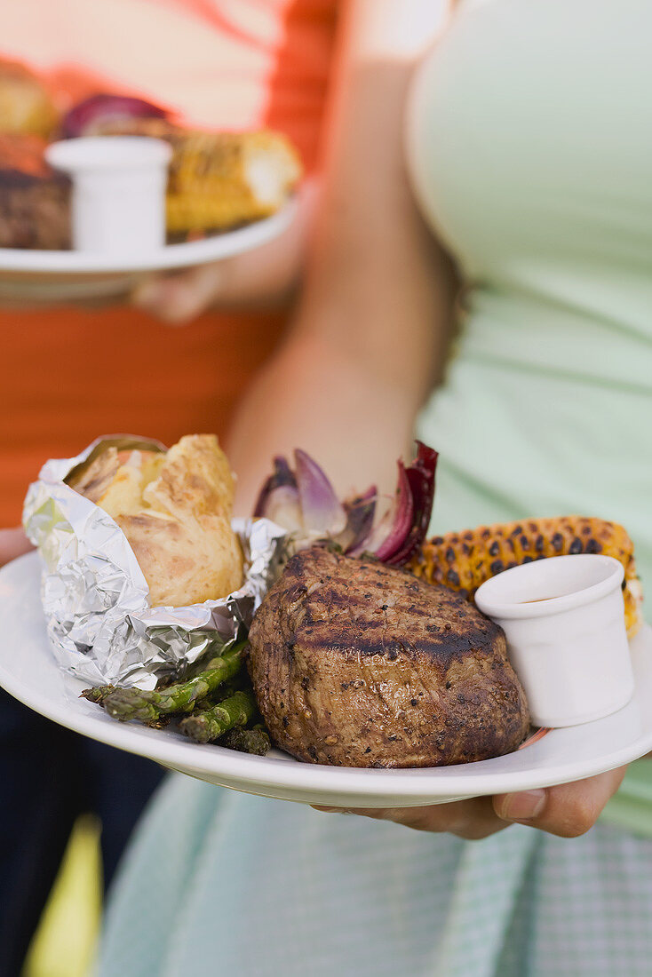 Two women holding plates of grilled steak & accompaniments