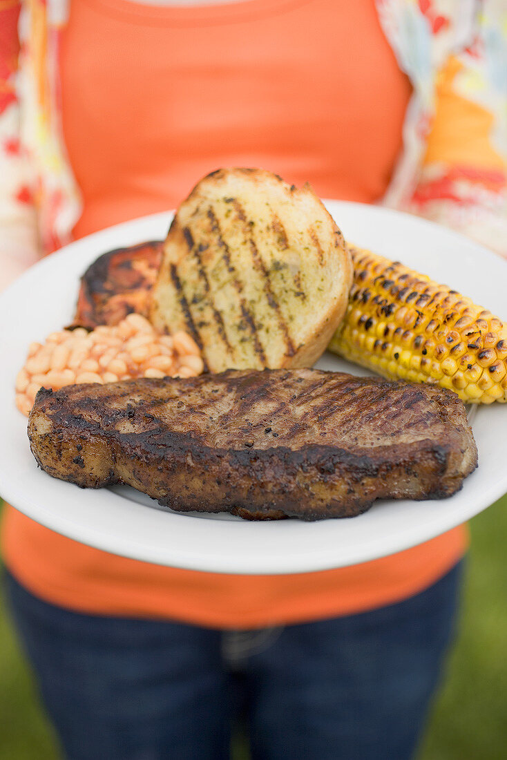 Woman holding plate of steak, bread, corn on the cob, baked beans