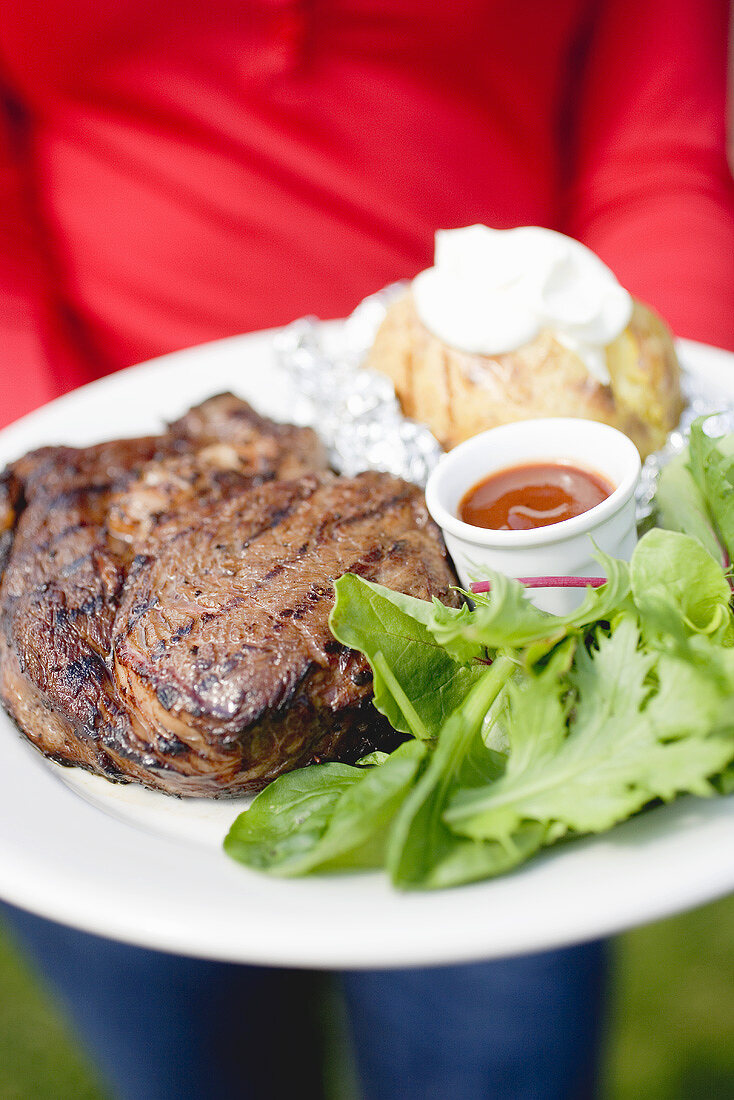 Person holding plate of grilled steak, baked potato & salad