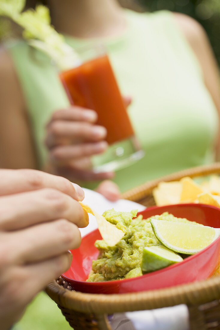 Hand holding basket of guacamole & chips, woman with tomato drink