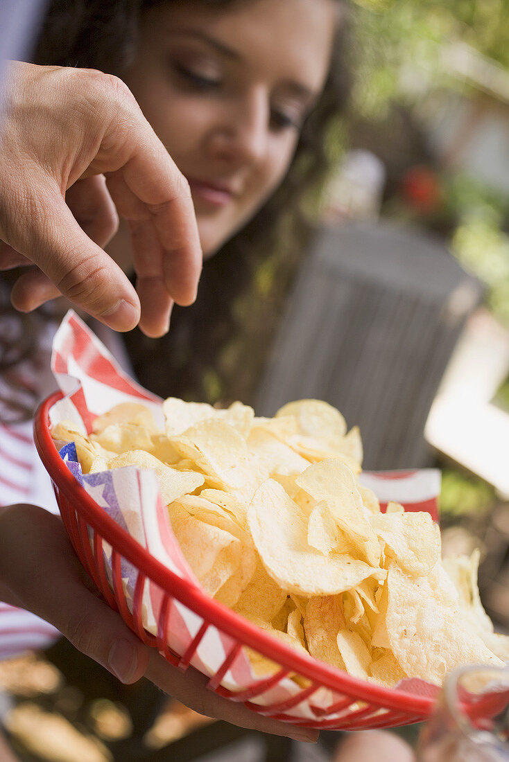 Hand reaching for crisps in plastic basket, woman in background