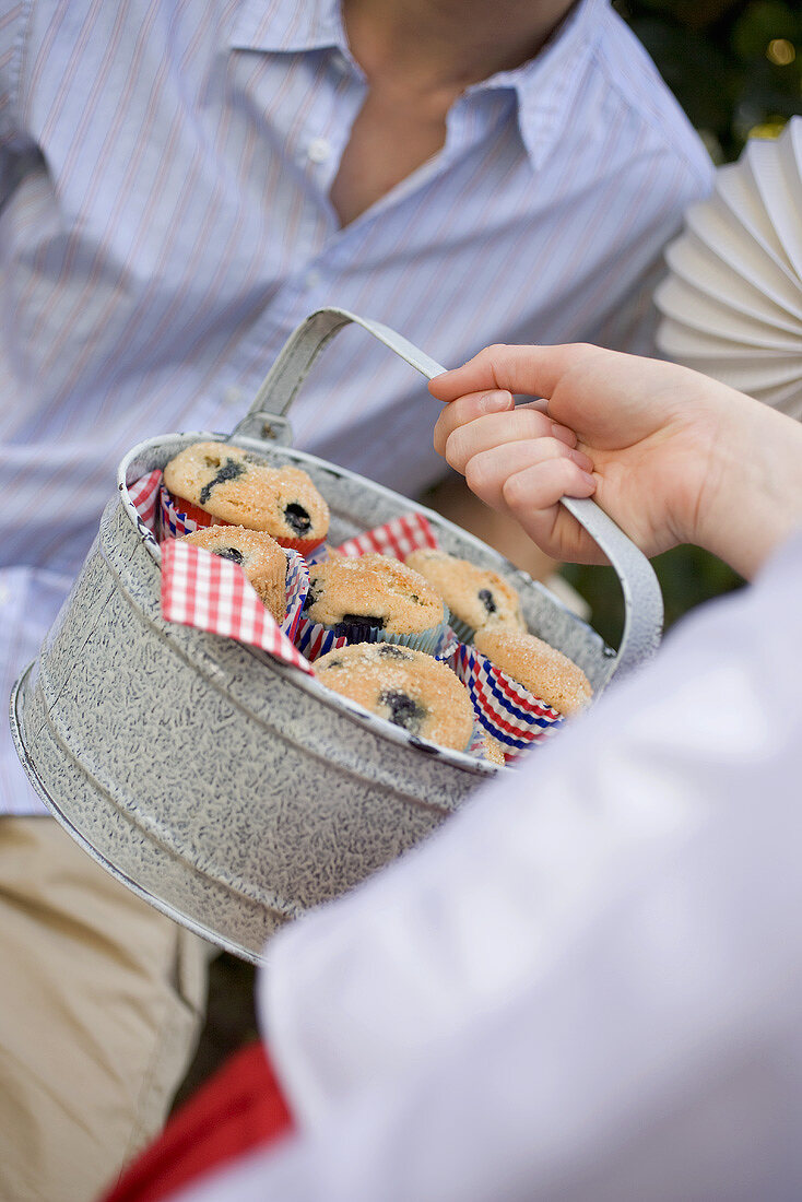 Hand holding a tin of blueberry muffins, man in background