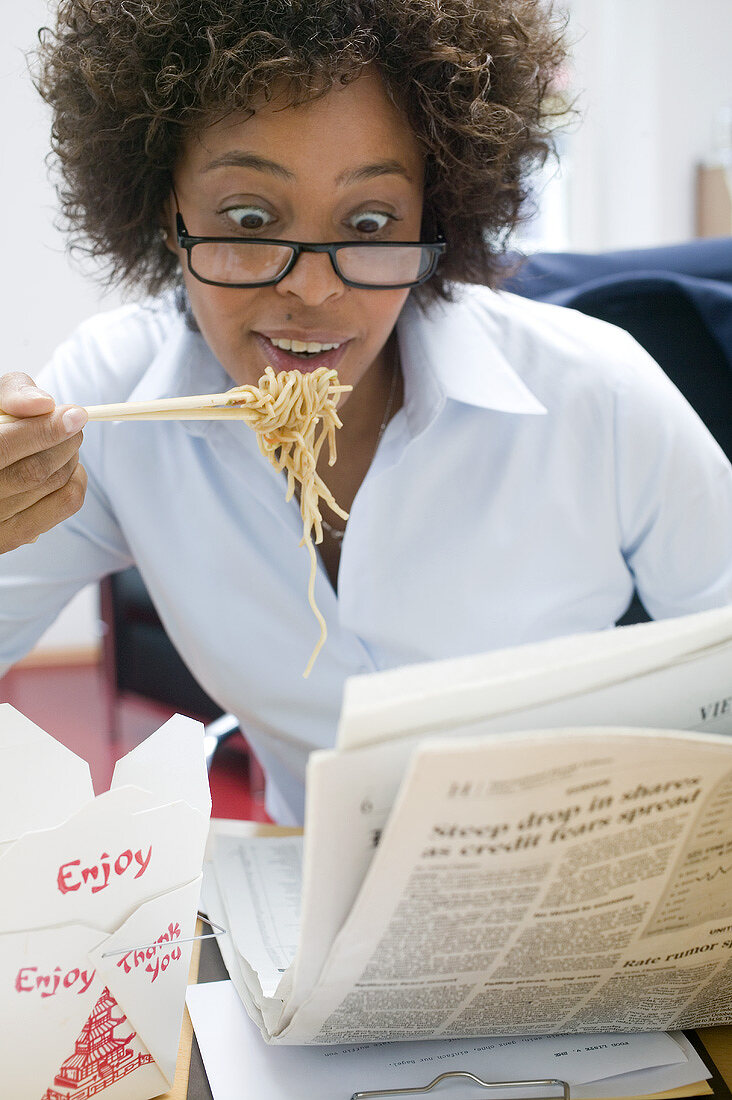 Frau im Büro isst asiatisches Nudelgericht beim Zeitunglesen
