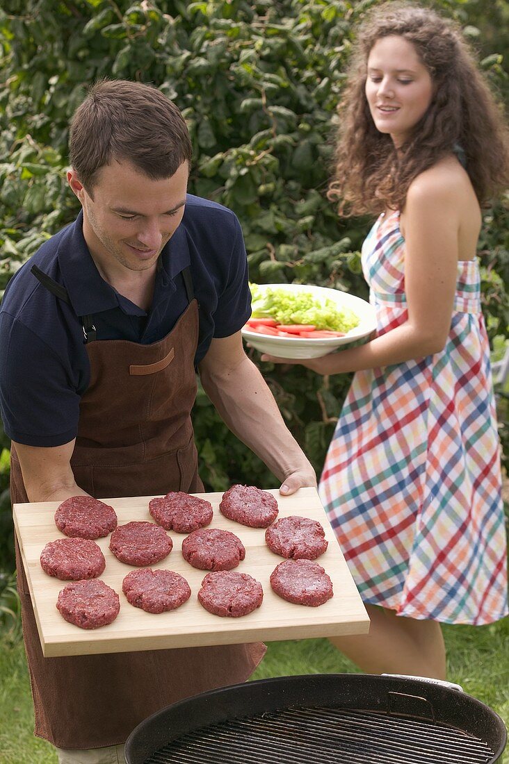 Man holding raw burgers over barbecue, woman serving salad
