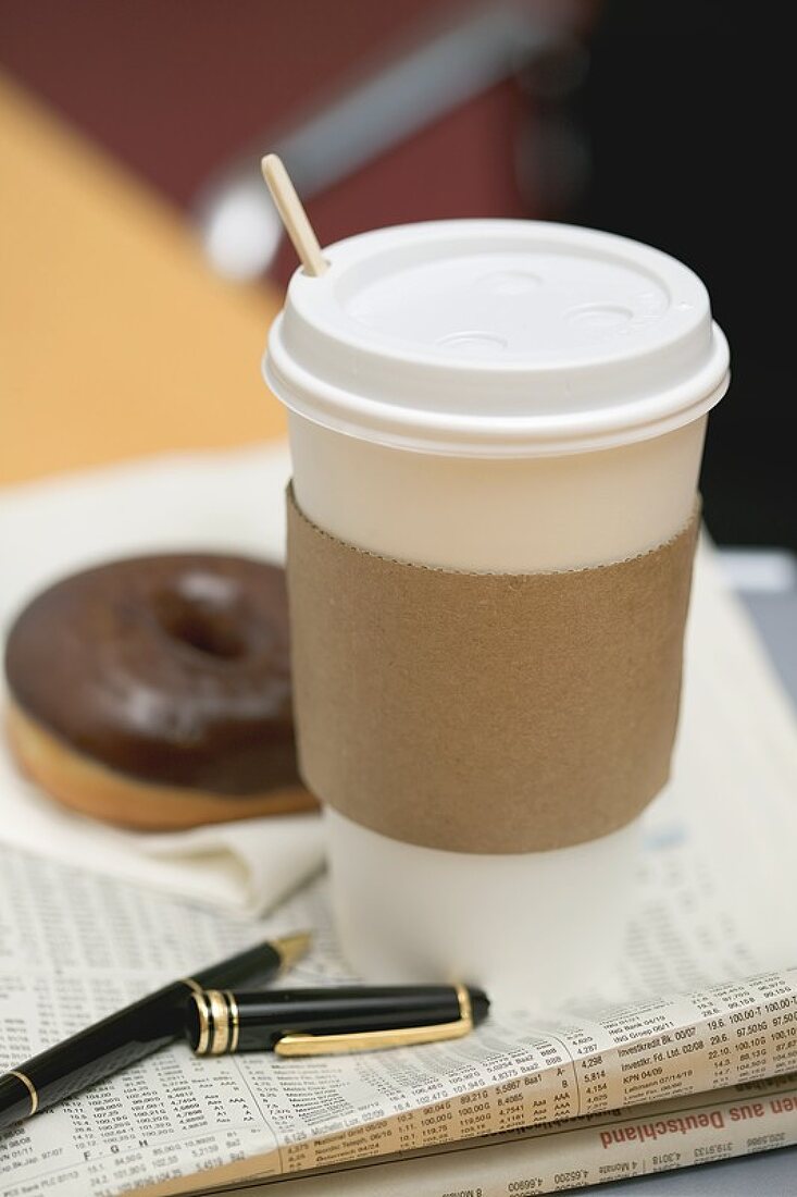 Coffee in paper cup and doughnut on newspaper in office
