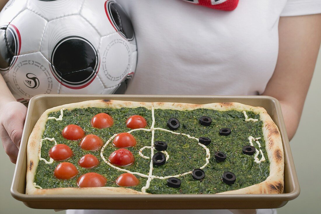 Fussballspielerin hält Spinatpizza mit Tomaten und Oliven