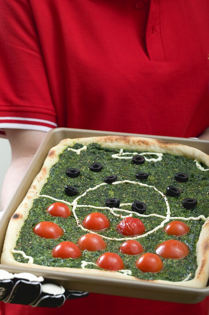 Fussballspielerin hält Spinatpizza mit Tomaten und Oliven