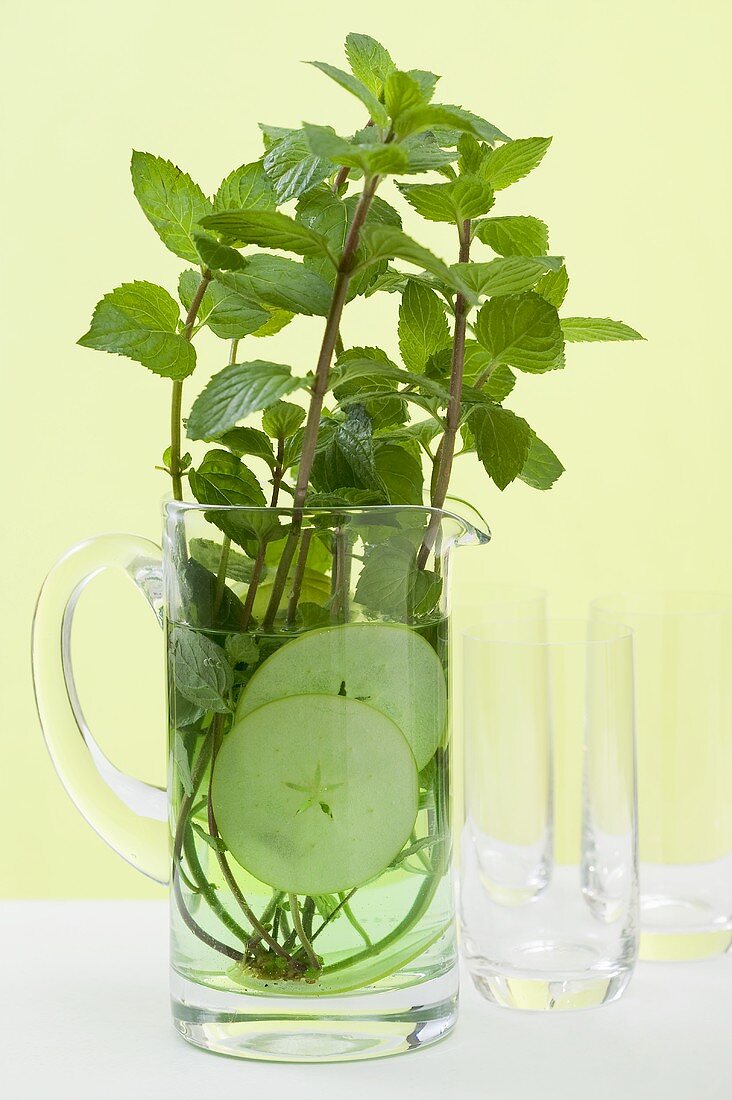 Mint sprigs & cucumber slices in jug of water in front of glasses