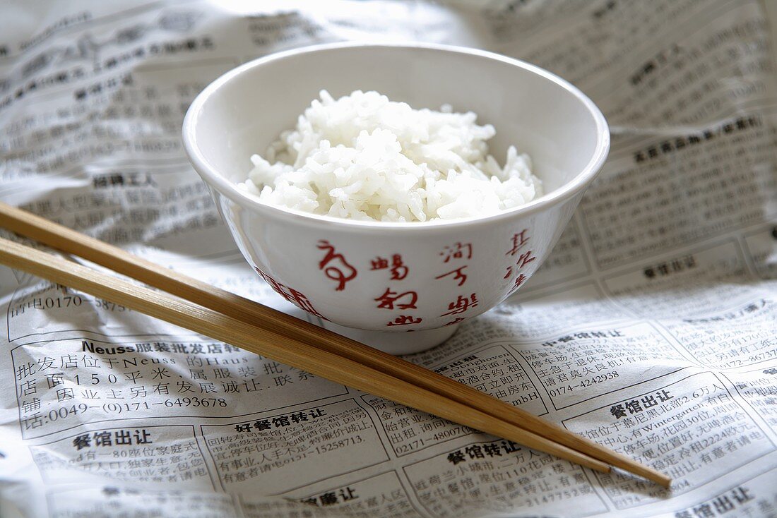 Reis in asiatischer Schale auf Zeitung, daneben Essstäbchen