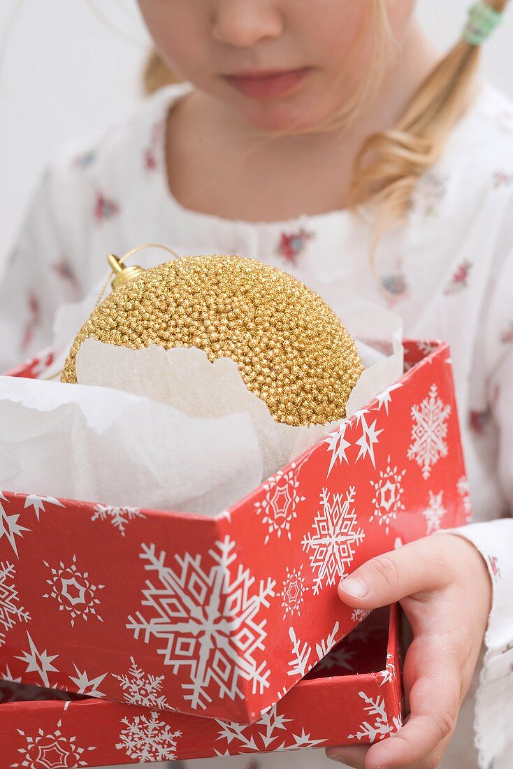 Kleines Mädchen hält Schachtel mit goldener Christbaumkugel