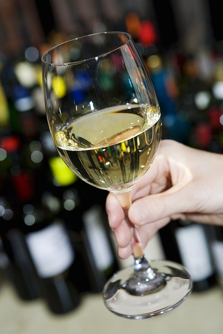 Hand hält ein Glas Weißwein, im Hintergrund Weinflaschen
