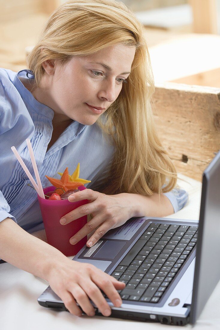 Blonde Frau mit Laptop und Cocktail auf dem Bett