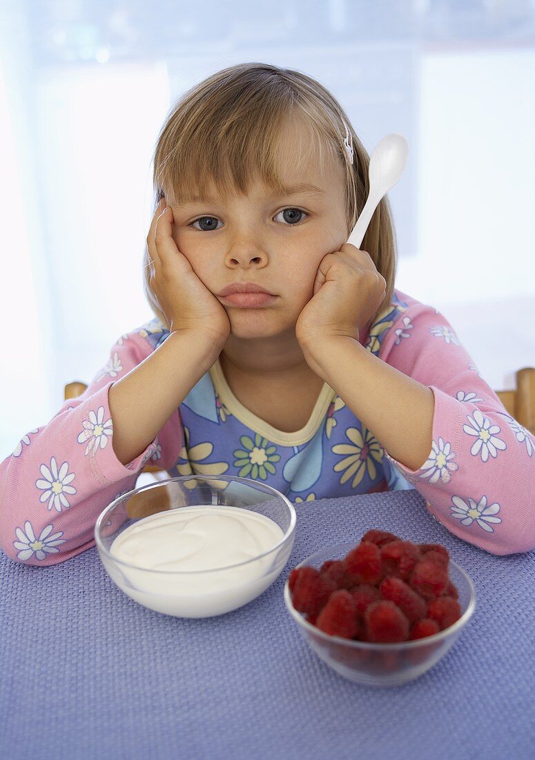 Mädchen sitzt lustlos vor Joghurt und Himbeeren