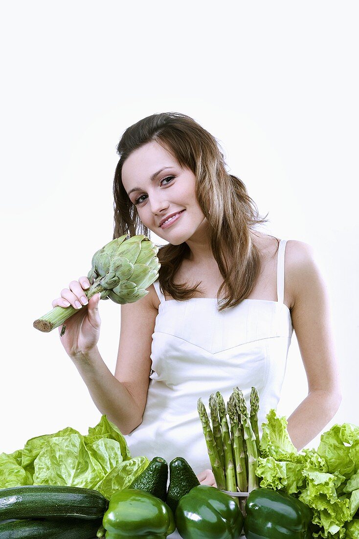 Junge Frau hält Artischocke in der Hand; grünes Gemüse davor