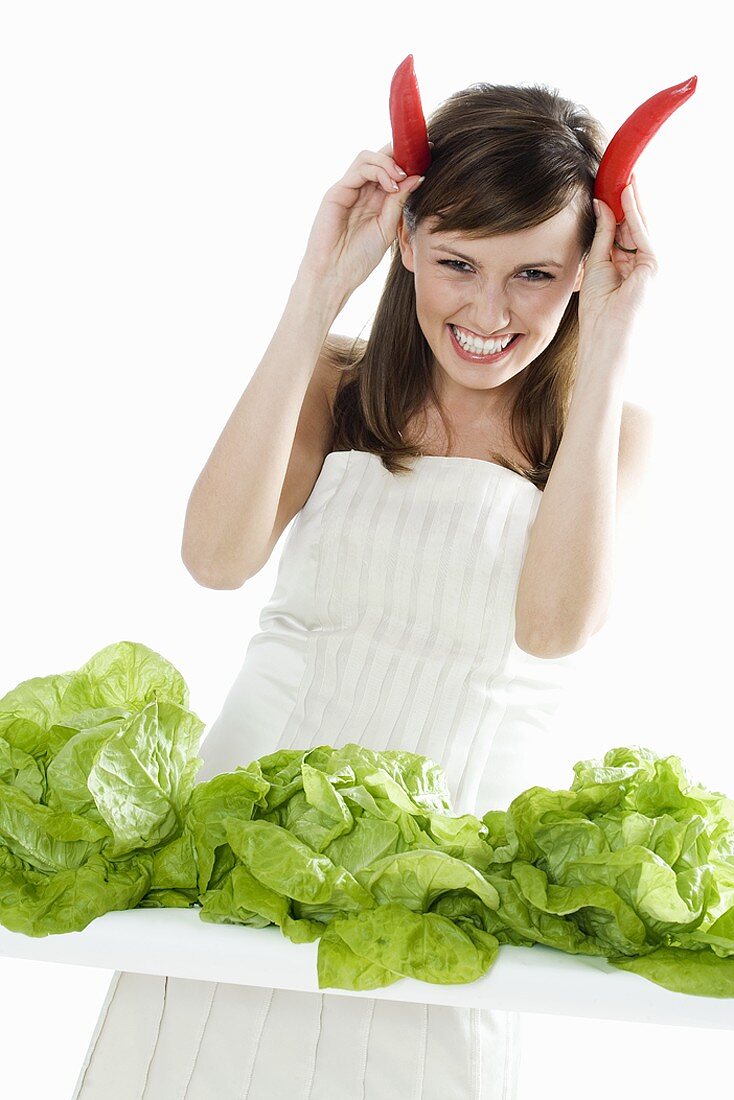 Junge Frau mit Peperoni-Hörnern; Kopfsalat davor