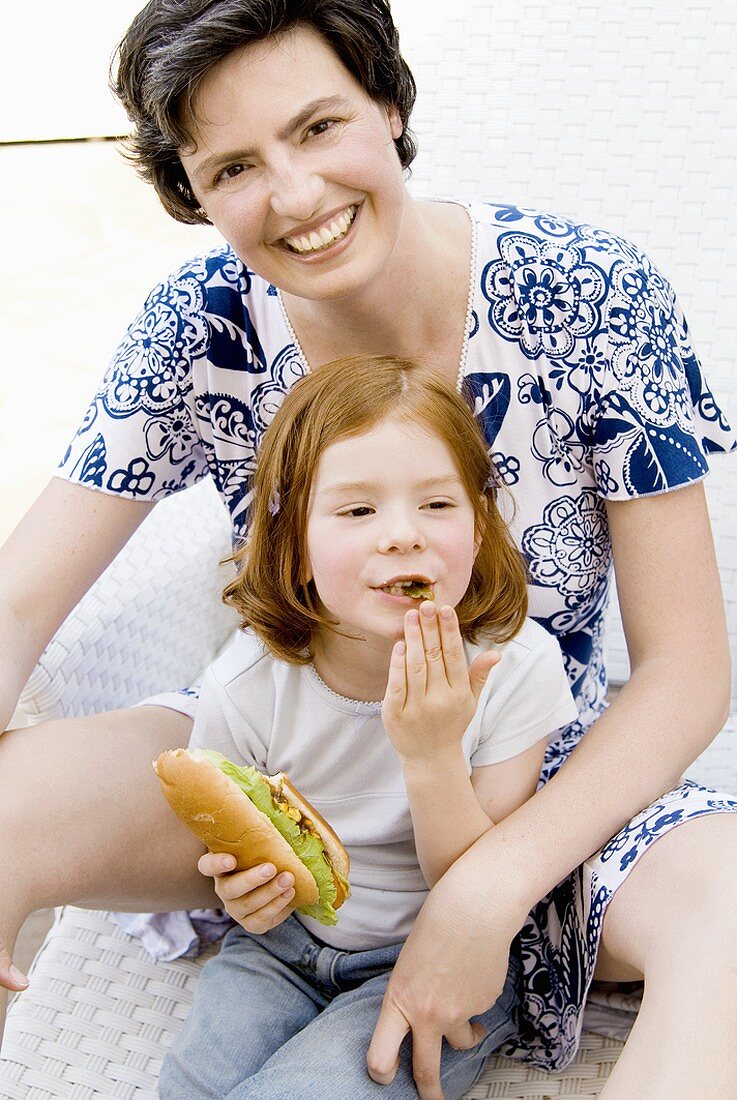 Frau sitzt hinter kleinem Mädchen mit Hot Dog