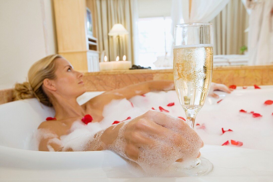 Frau mit Glas Sekt in Badewanne nimmt Rosenblütenblätter-Bad