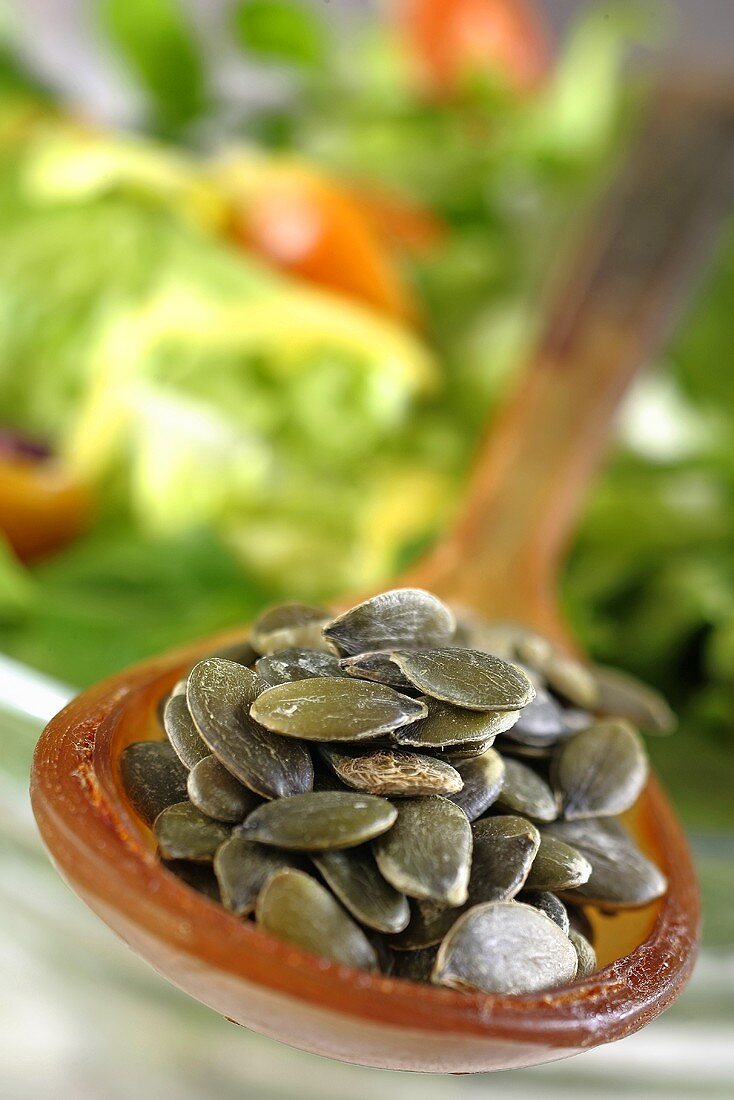 Pumpkin seeds on a spoon in from of a bowl of salad