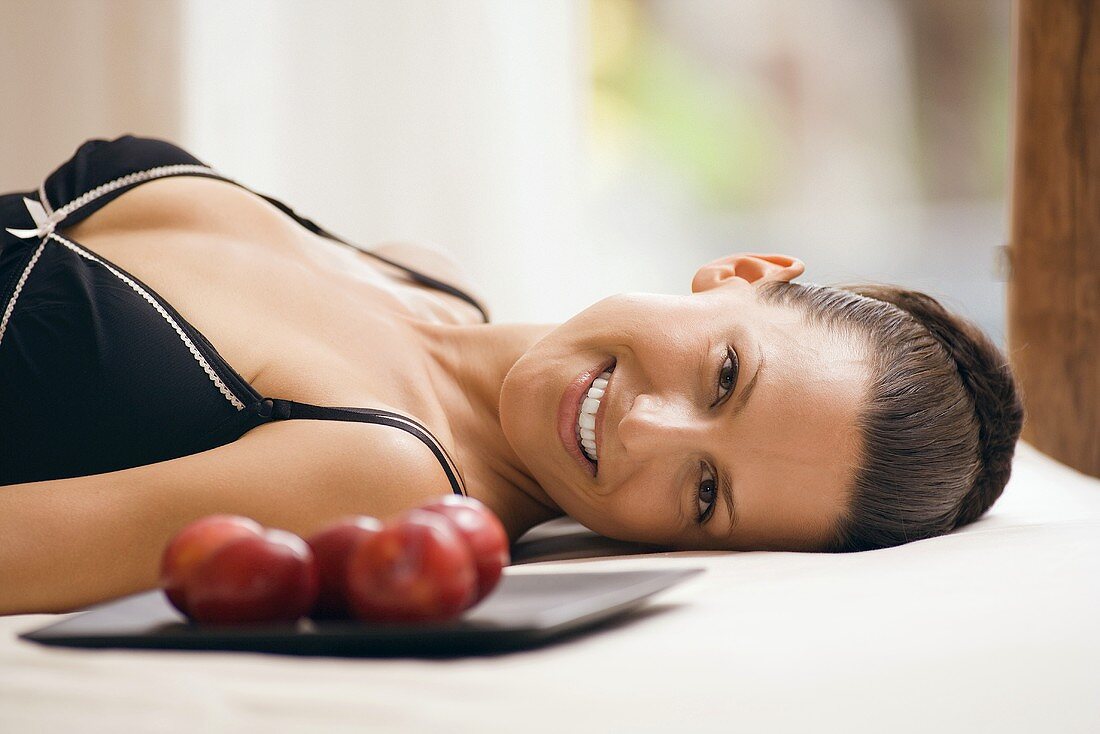 Young woman wearing neglige, relaxing on bed alongside tray with plums