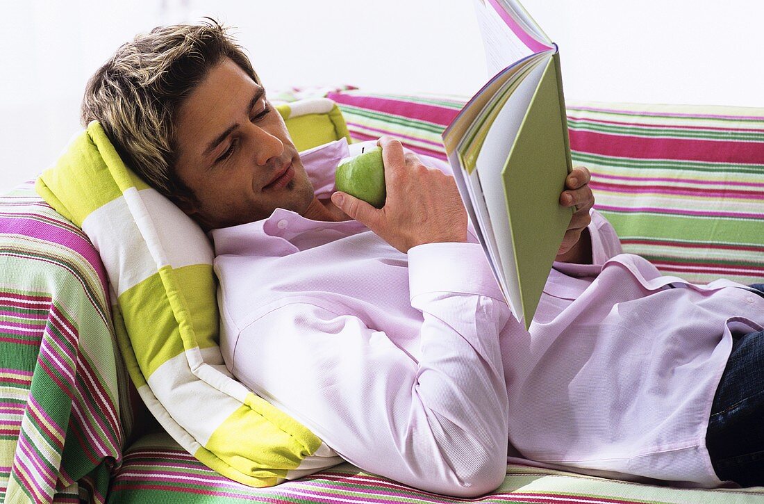Young man lying on sofa, reading book, side view