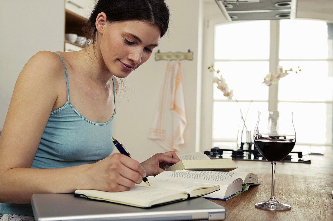 Young woman sitting at table with books, laptop and red wine