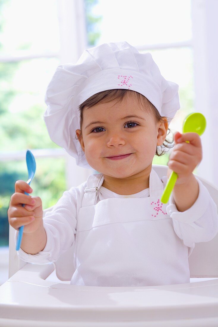 Kleines Mädchen mit Kochmütze hält Plastiklöffel in der Hand