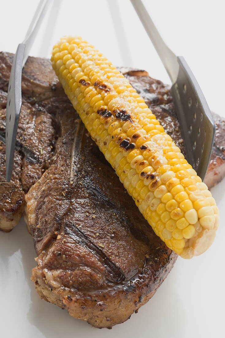 Grilled T-bone steak with corn on the cob and grill tongs