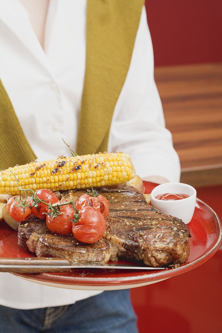 Woman holding T-bone steak, corn on the cob & cherry tomatoes