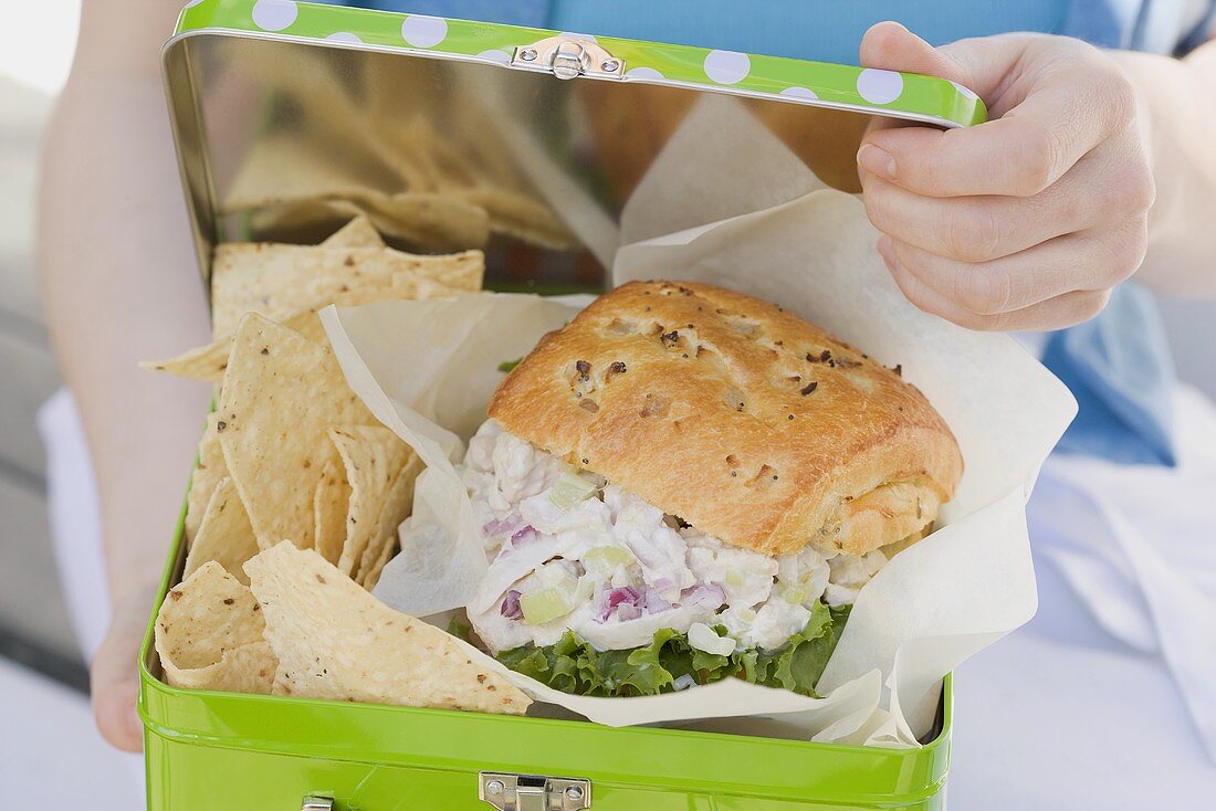 Woman holding lunch box containing chicken sandwich & crisps