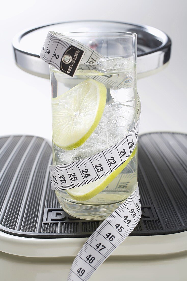 Glass of water with lime & tape measure on scales (close-up)
