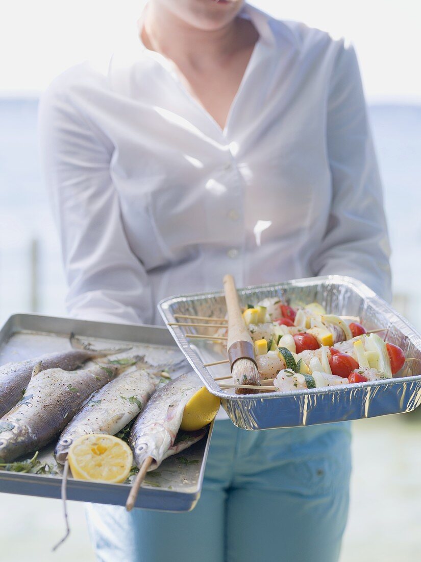 Woman holding trays of fish & kebabs ready for grilling, outdoors