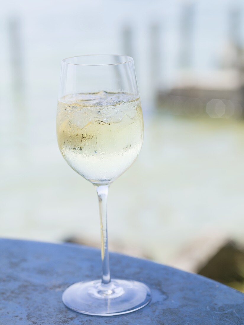 Weissweinglas mit Eiswürfeln auf Tisch am Seeufer