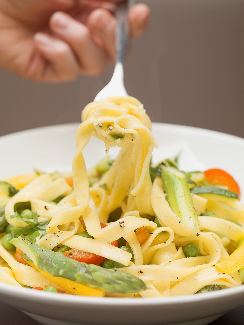 Hand holding a forkful of ribbon pasta and vegetables