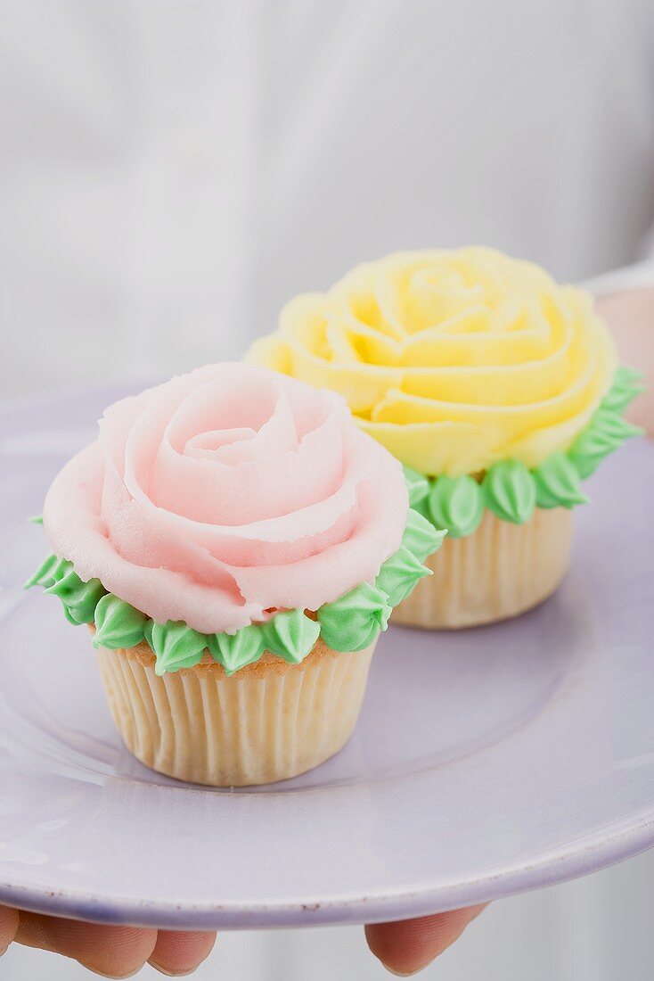 Hands holding two cupcakes with marzipan roses on plate