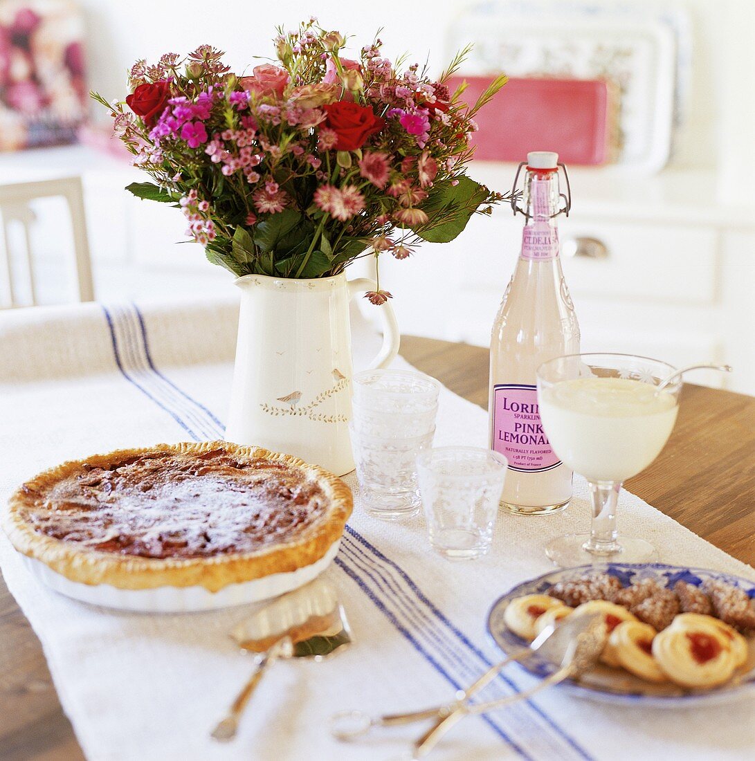 Tart, small cakes, lemonade and jug of flowers on table